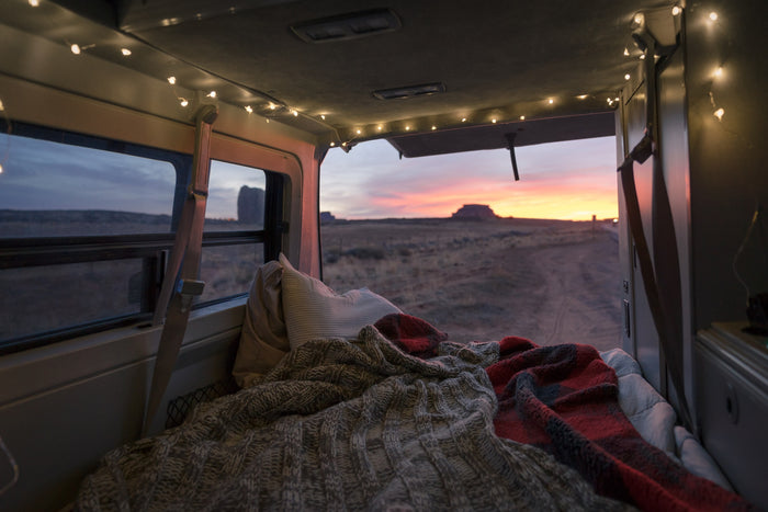 Experience cozy RV rental living: van interior with string lights, blankets, and a bed, overlooking a stunning desert sunset & rock formations.