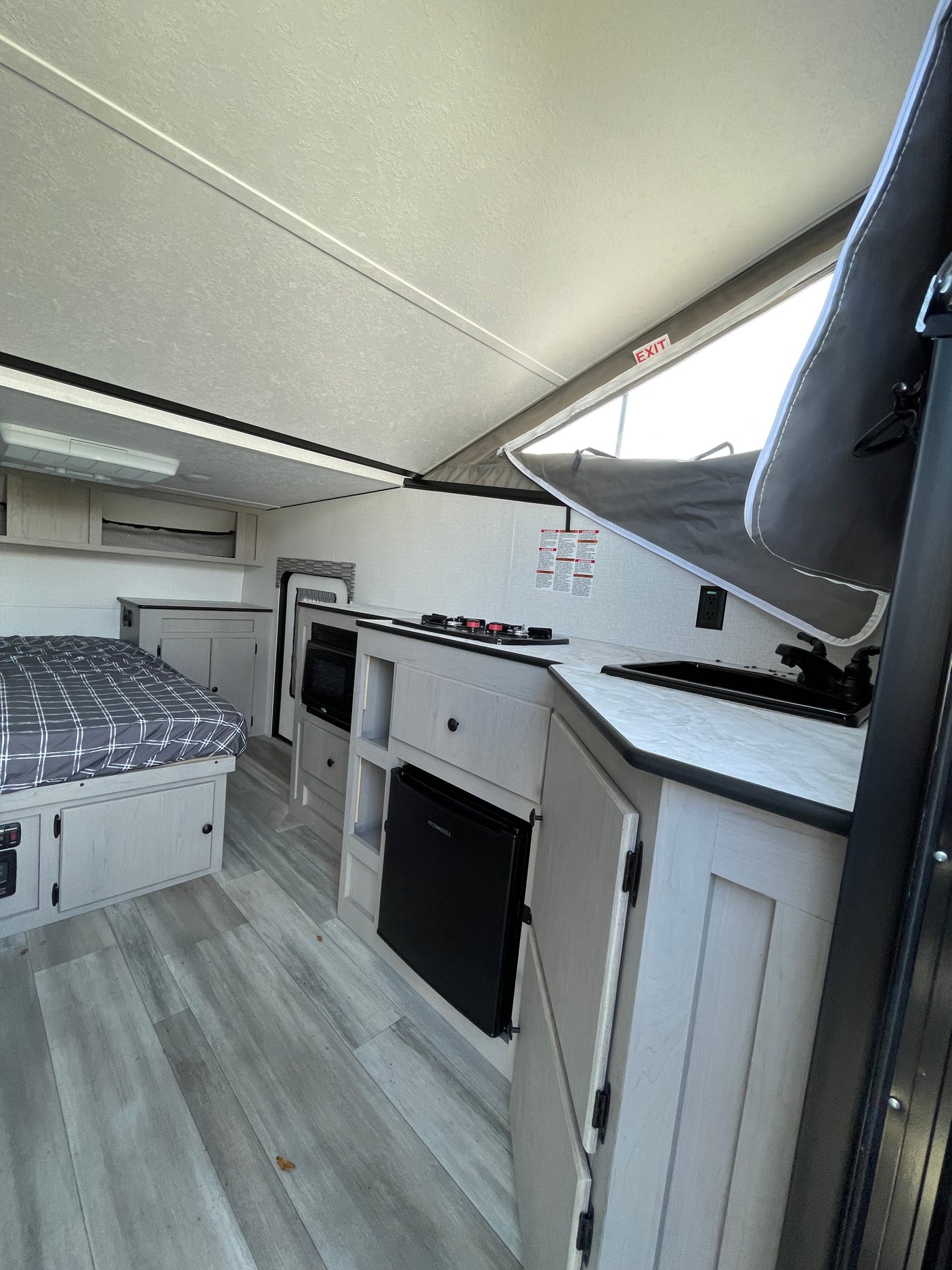 Bright rv rental interior featuring a bed, kitchenette with stove, sink, microwave, mini-fridge, and pop-up roof. Light wood finishes.