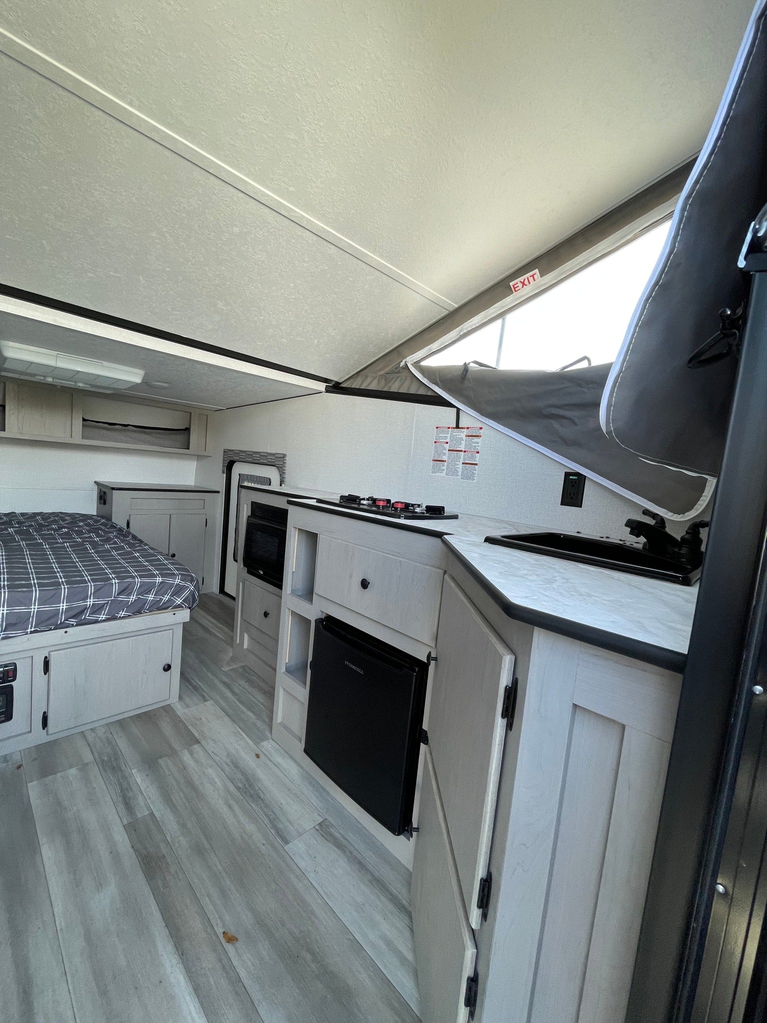 Bright rv rental interior featuring a bed, kitchenette with stove, sink, microwave, mini-fridge, and pop-up roof. Light wood finishes.