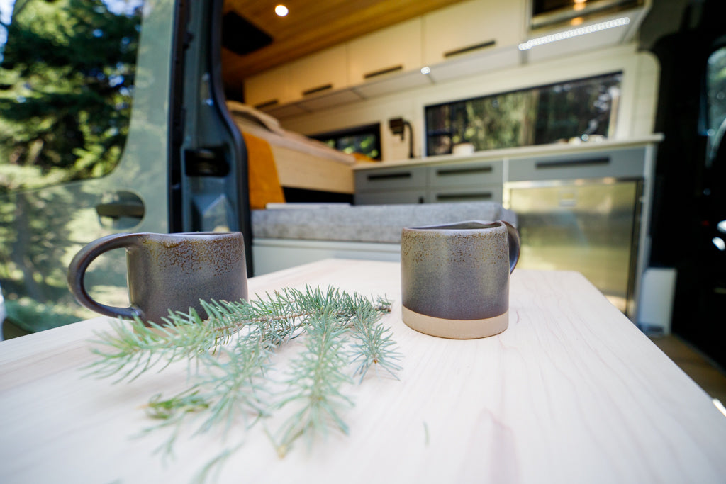 Enjoy nature from your RV rental! Two ceramic mugs and evergreen sprigs on a wood table inside a modern camper van.