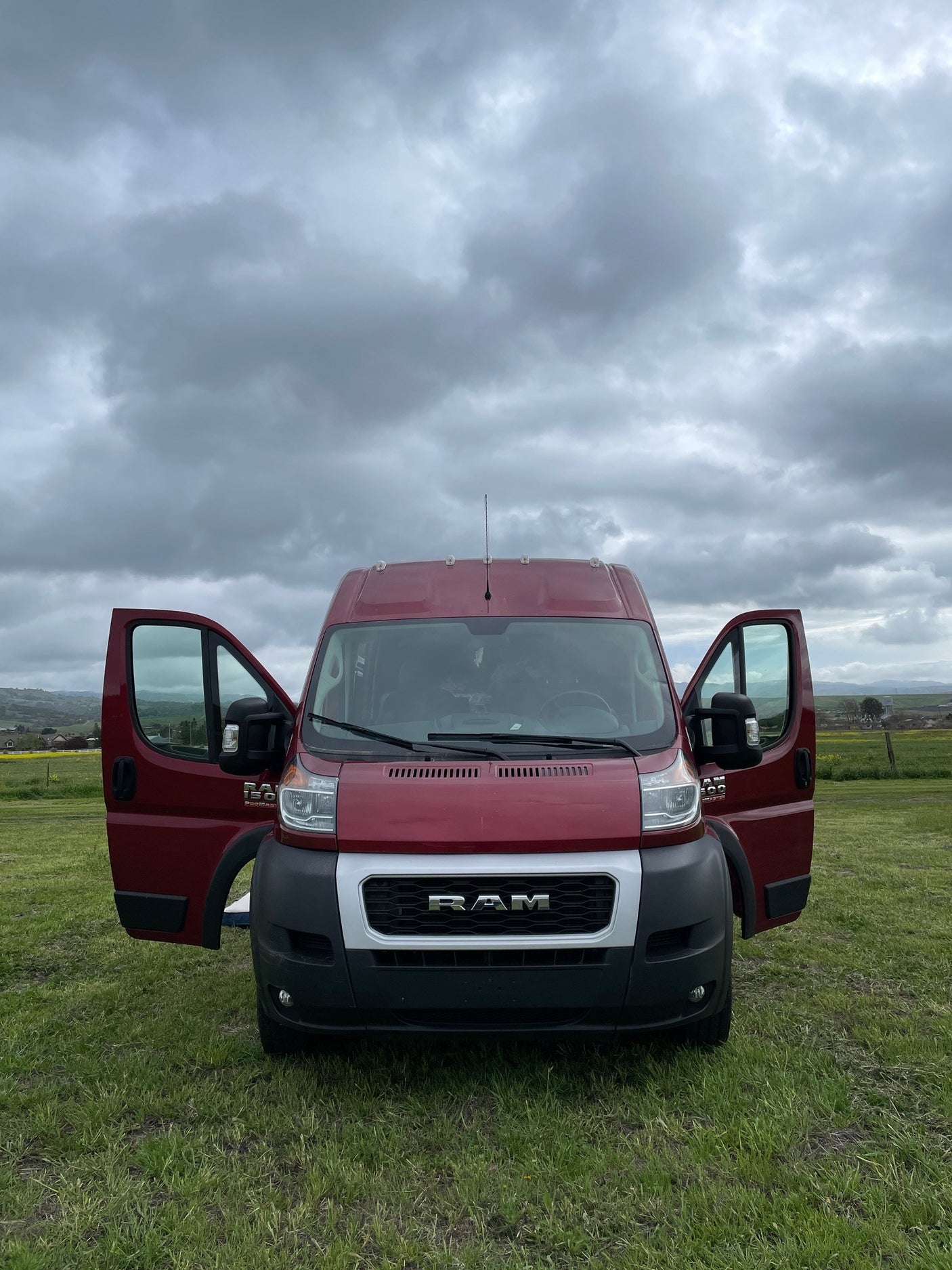 Red Ram ProMaster van for RV rental, doors open in a green field under cloudy skies. Your travel adventure starts here!