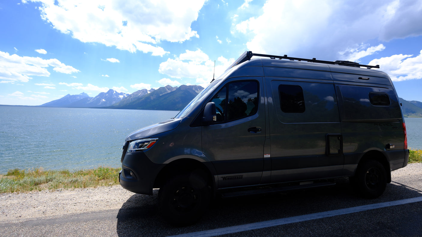 Gray Winnebago Revel rv rental parked beside a large lake with distant mountains under a blue sky with white clouds.