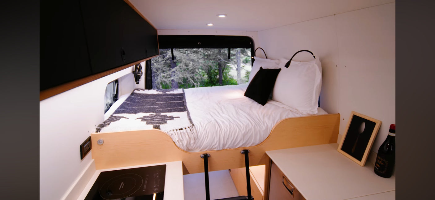 RV rental interior: cozy bed with patterned blanket, compact kitchen, and storage. Outside, lush trees are visible.