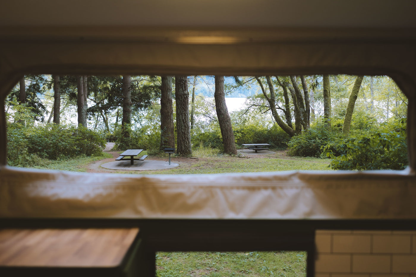 Scenic RV rental view of a peaceful forest campsite with picnic tables and a distant lake, seen from inside the vehicle. — photo 13