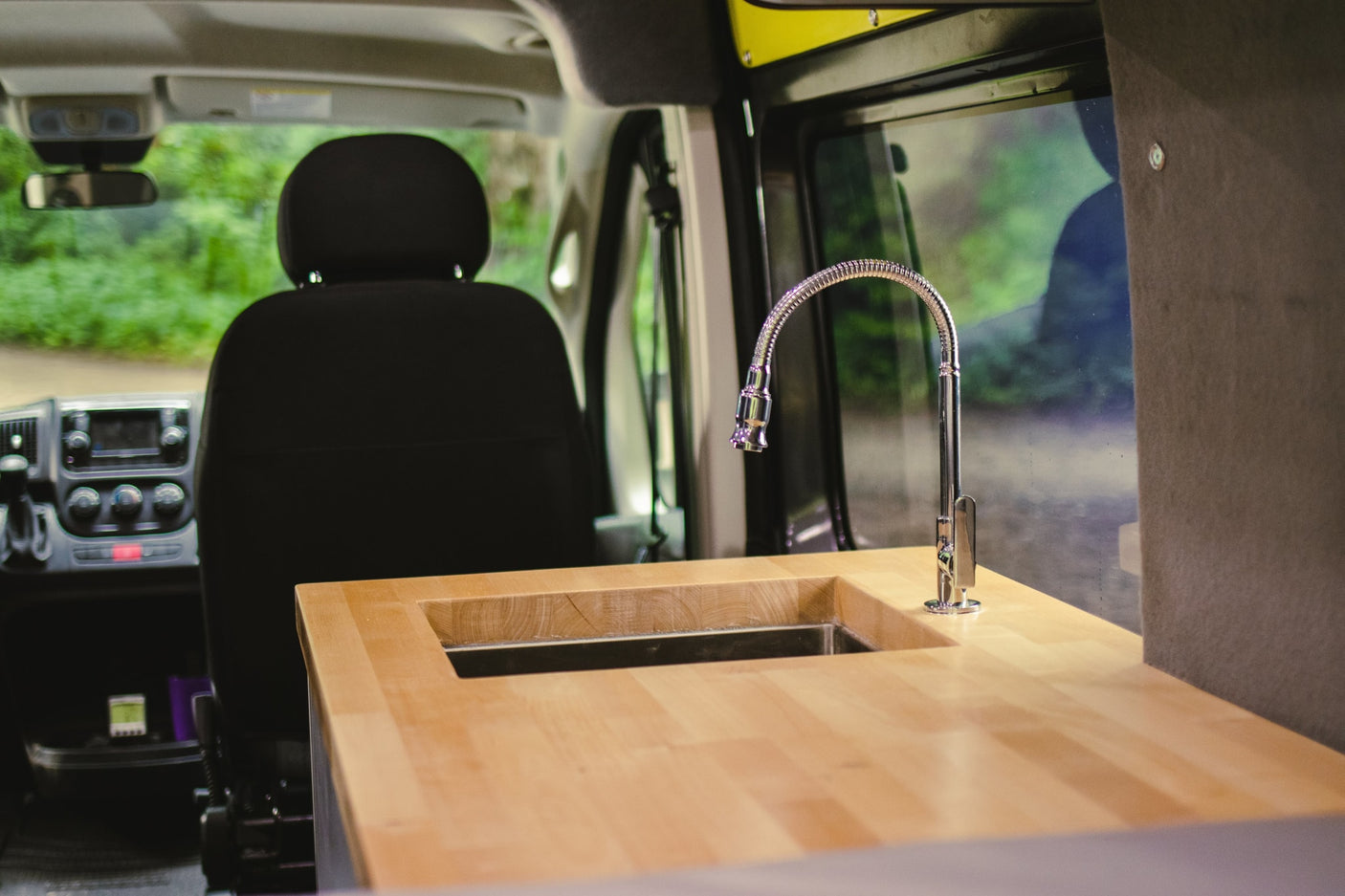 An RV rental's van conversion kitchen: wooden counter, integrated sink, and flexible faucet, with lush greenery outside.