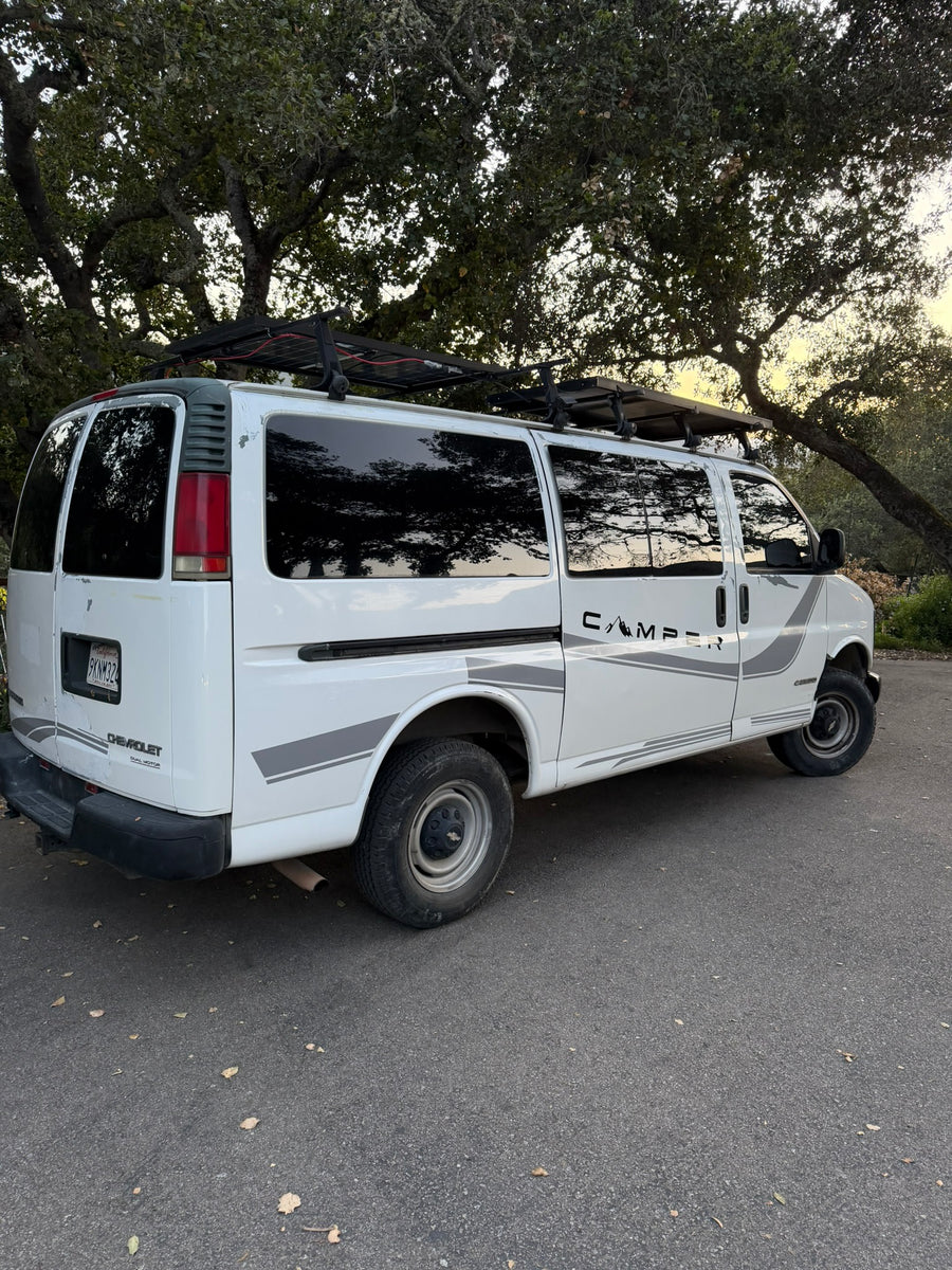 White Chevy Express camper van with rooftop solar panels and "CAMPER" decal, parked under trees. Ready for your rv rental adventure.