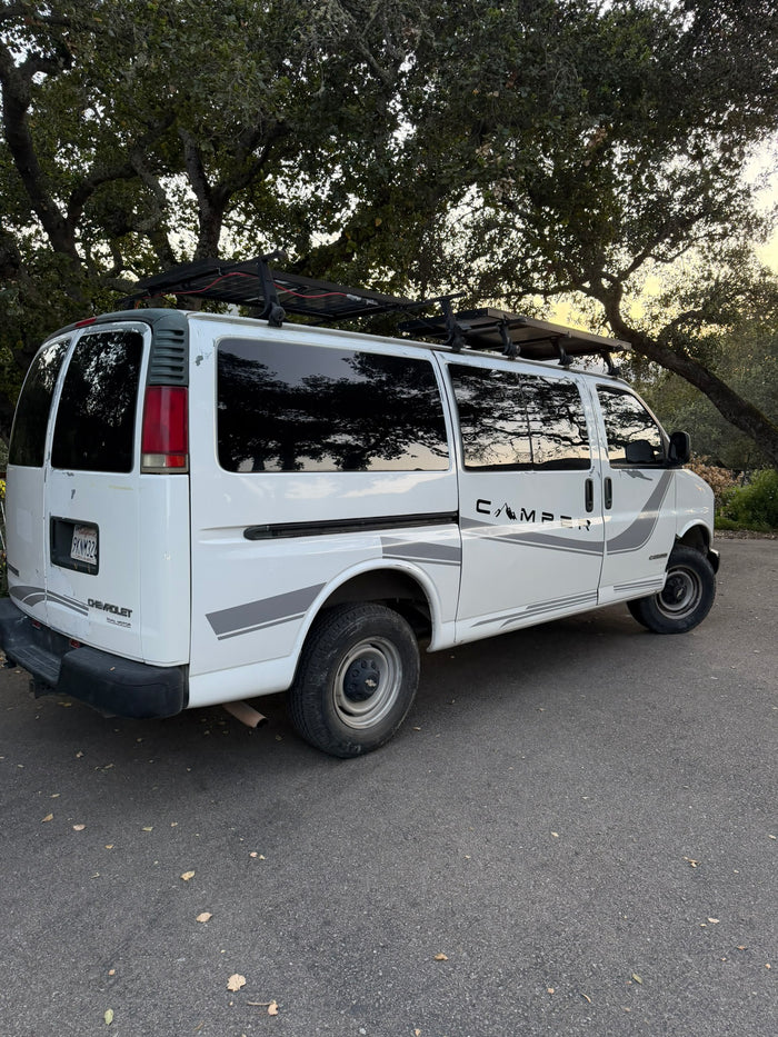 White Chevy Express camper van with rooftop solar panels and "CAMPER" decal, parked under trees. Ready for your rv rental adventure.