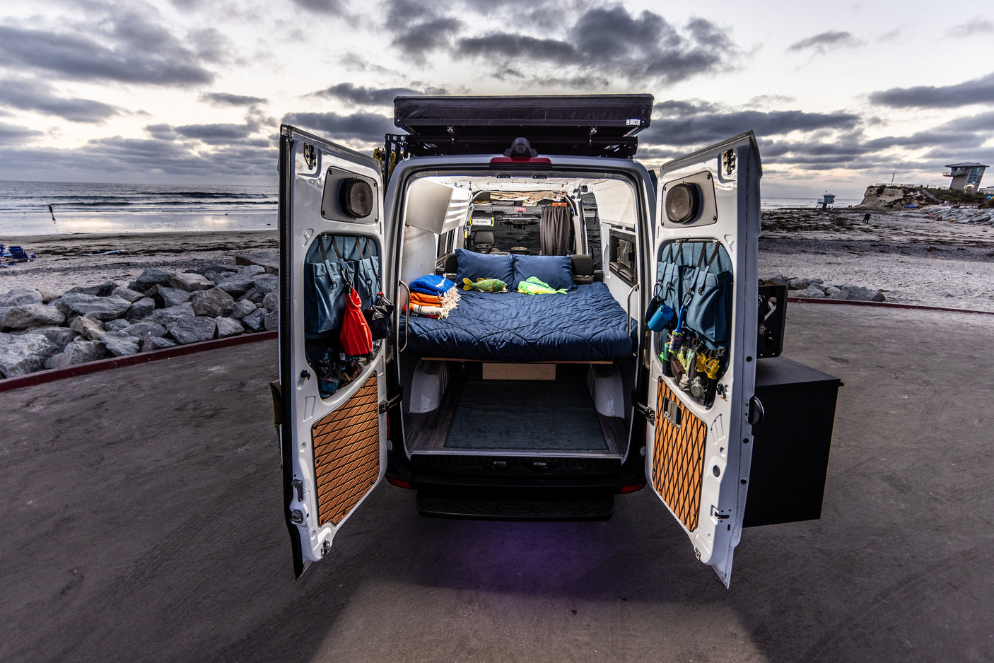 This RV rental camper van, parked beachside, reveals a cozy bed and organized interior through its open rear doors.
