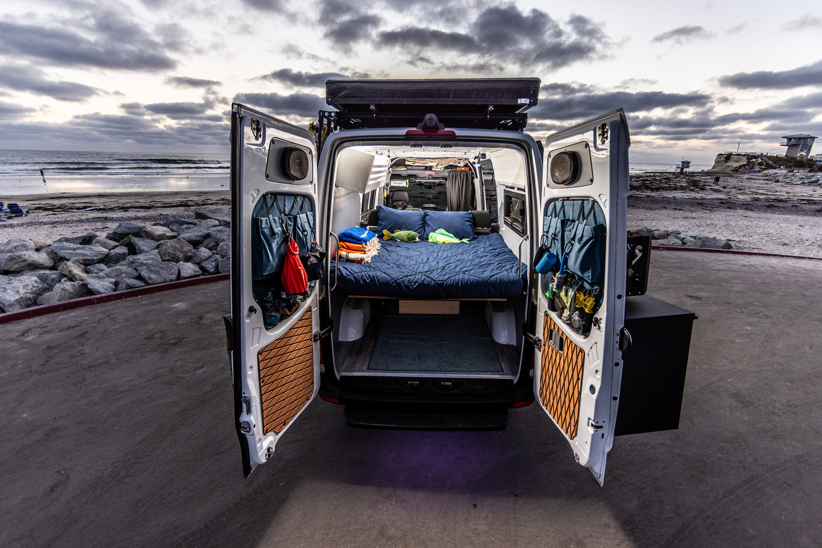 This RV rental camper van, parked beachside, reveals a cozy bed and organized interior through its open rear doors.