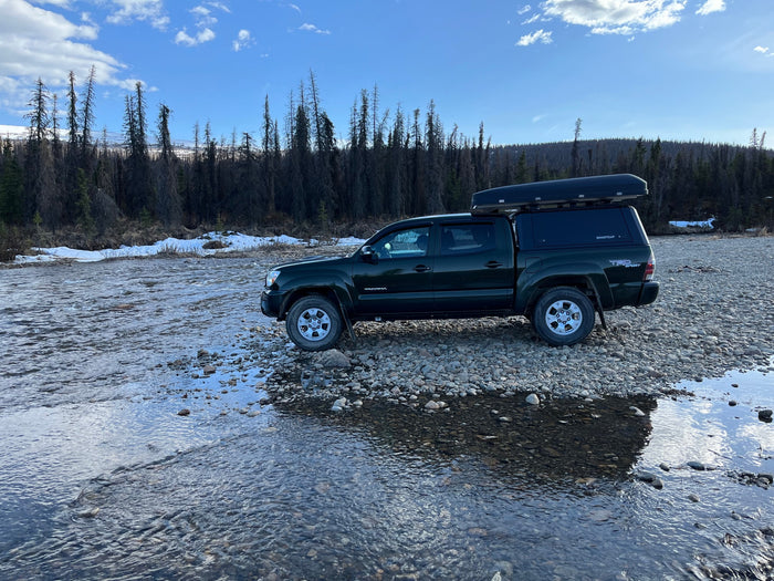 Dark green truck with rooftop tent on a rocky riverbed, perfect for an off-grid RV rental adventure. Mountains & forest view.