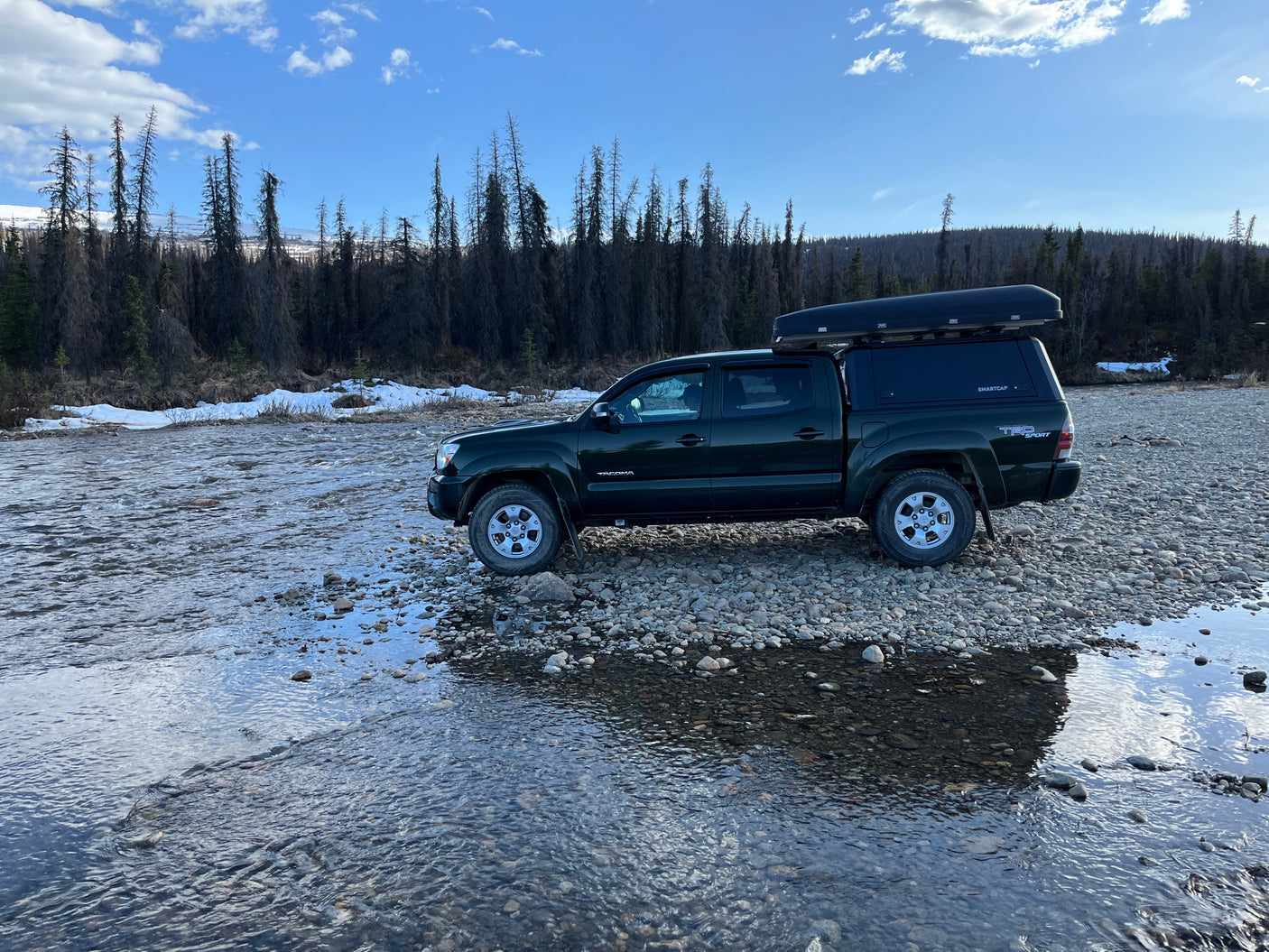 Dark green truck with rooftop tent on a rocky riverbed, perfect for an off-grid RV rental adventure. Mountains & forest view.