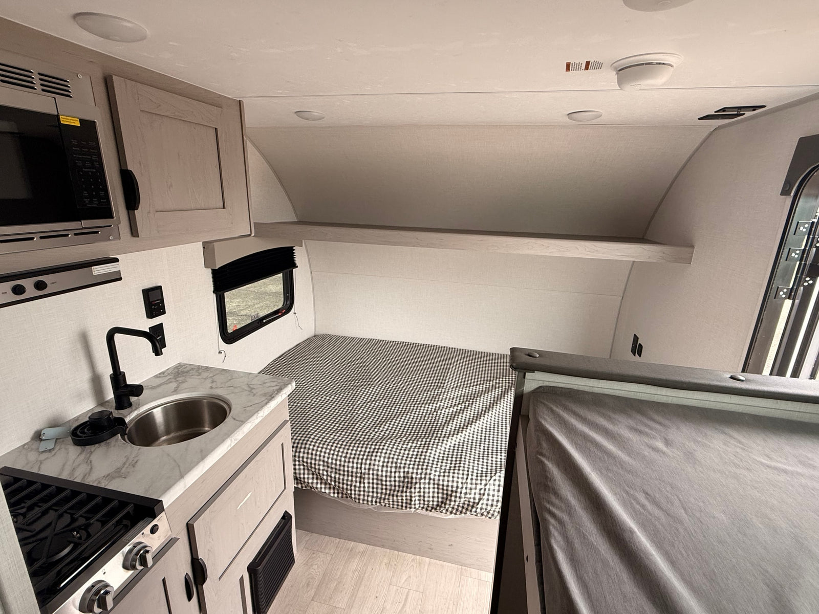 This rv rental features a compact interior with a kitchen (sink, stove, microwave), a bed with gingham bedding, and overhead storage.