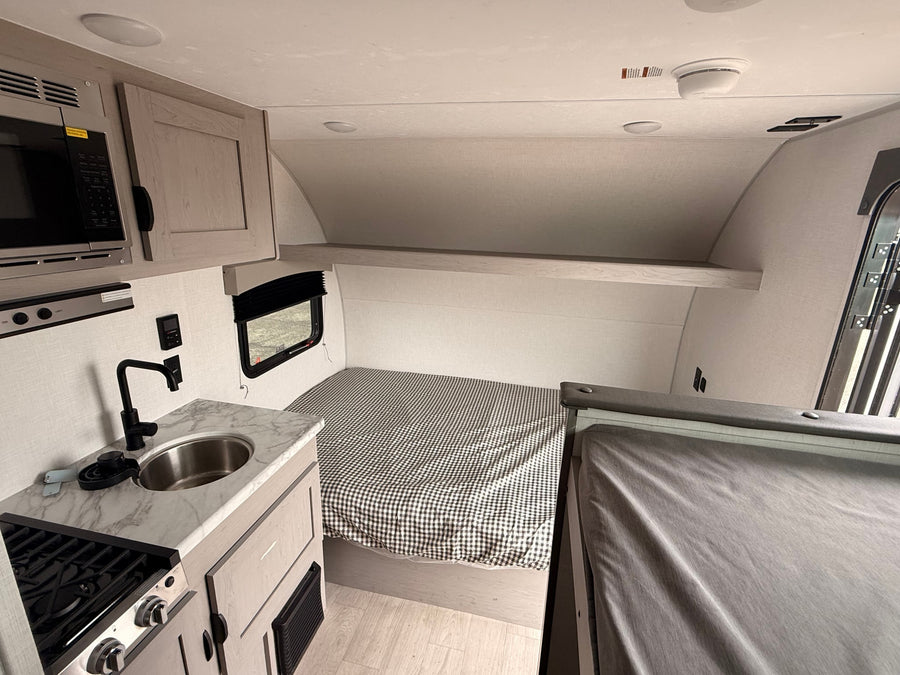 This rv rental features a compact interior with a kitchen (sink, stove, microwave), a bed with gingham bedding, and overhead storage.