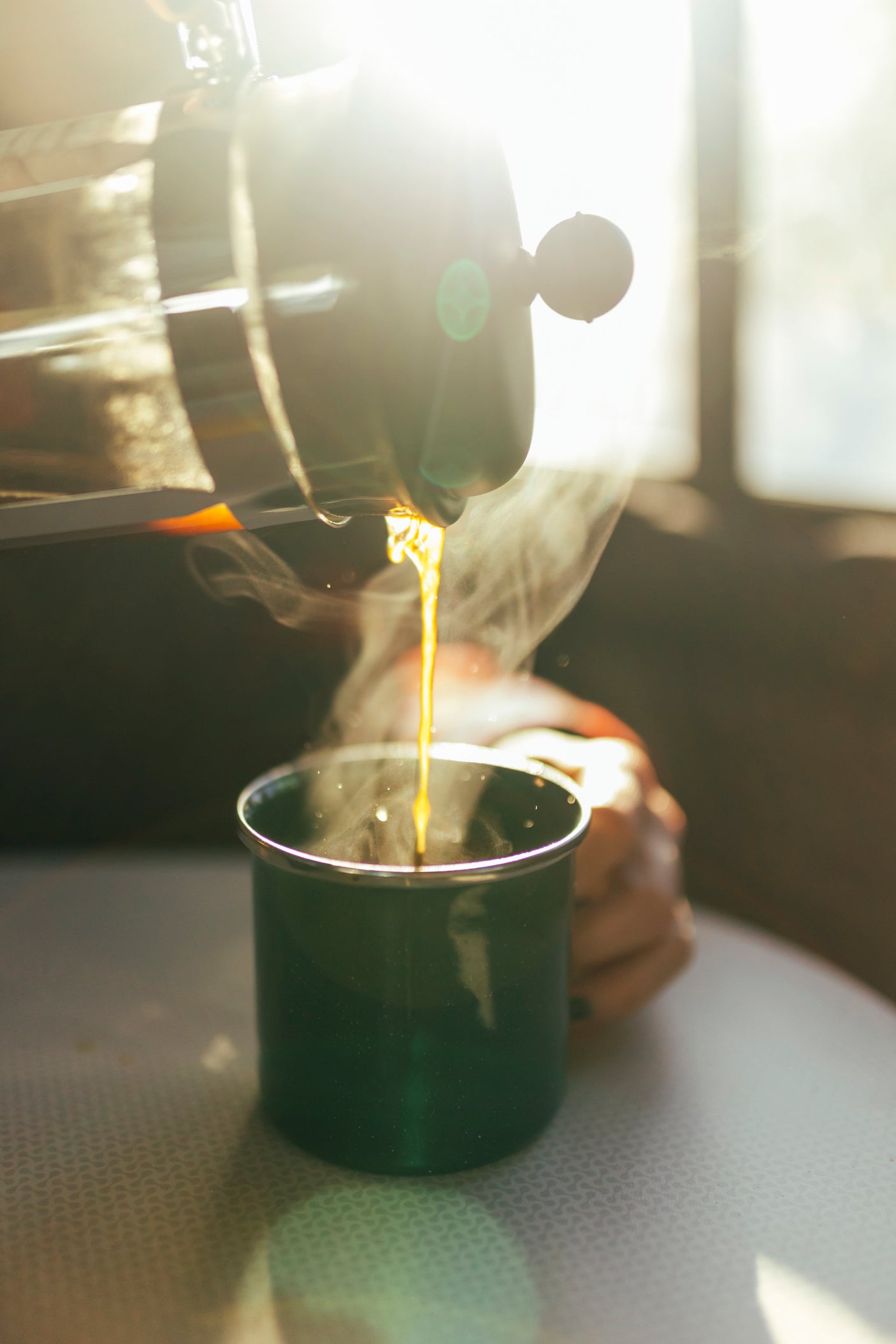 Person pouring steaming coffee from a French press into a green mug, bright morning inside an RV rental.