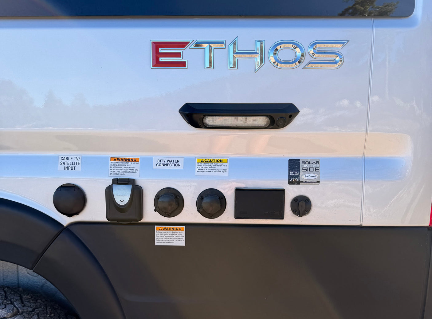 Ethos RV rental's utility panel, showing inputs for cable/sat, city water, shore power, and solar hookups, plus safety warnings.