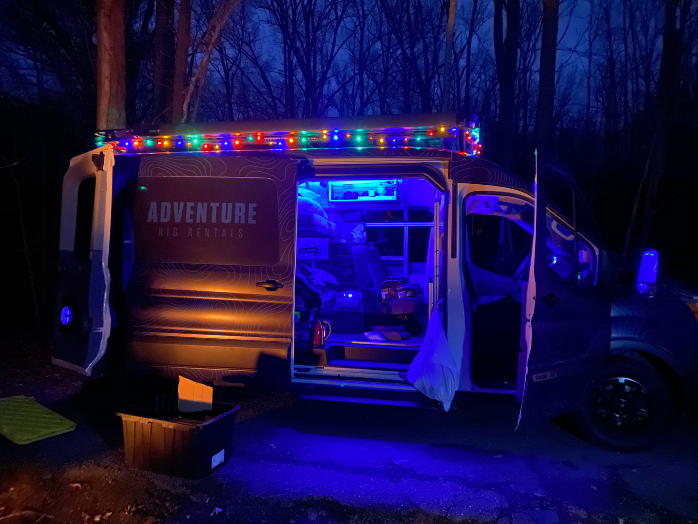 Adventure Rig RV rental van at night, illuminated by colorful string lights and a bright blue interior, parked for a cozy camp.