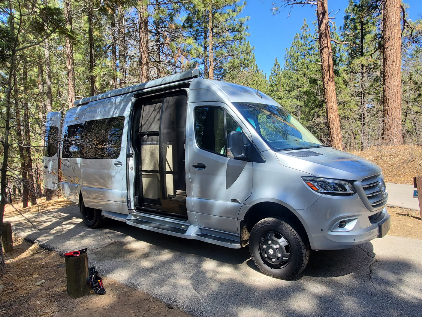 Silver Mercedes Sprinter RV rental parked in a sunny forest campsite, side door open. Shoes on a stump nearby.
