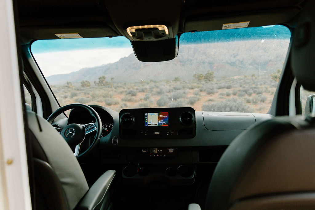 Inside a Mercedes-Benz rv rental van, the dash and screen show a scenic desert road trip view with mountains in the distance.