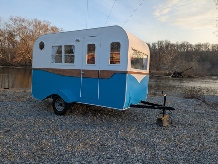 Unique blue & white RV rental, a compact camper parked by a serene river on a gravel bank for your next adventure.
