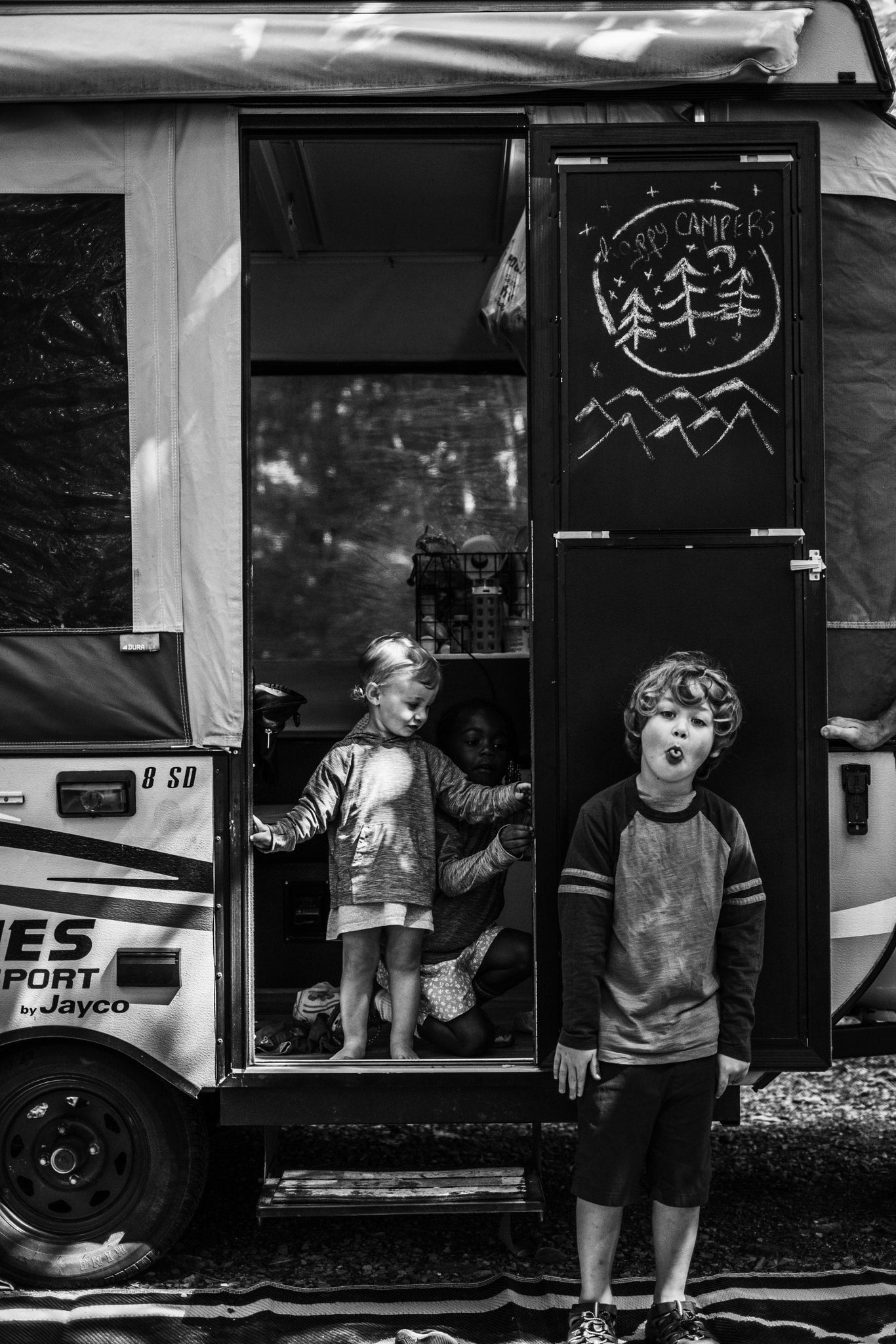An RV rental brings joy: Kids play around a pop-up camper, with one boy making a silly face outside. "Happy Campers" chalkboard visible.