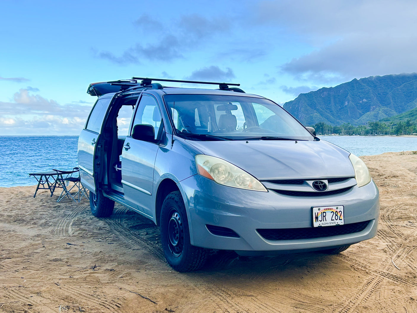 A grey minivan RV rental on a sandy Hawaii beach, door open, camping gear visible. Ocean and mountains in the background.