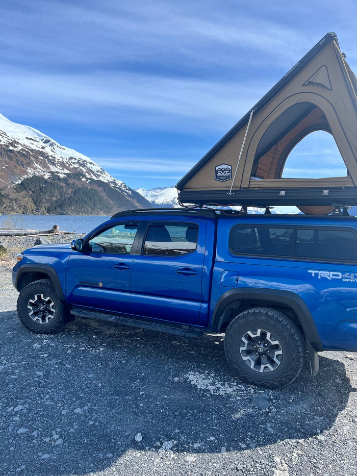 Adventure-ready blue pickup with rooftop tent on a lake shore, snow-capped mountains in back. Consider this for an RV rental trip.