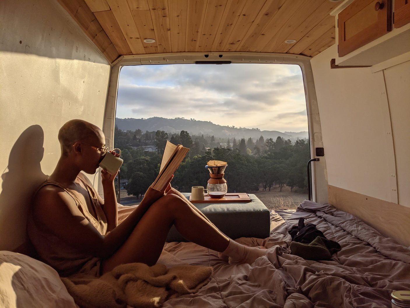 A person enjoys coffee & a book in an rv rental, with open back doors showing misty mountains & trees.
