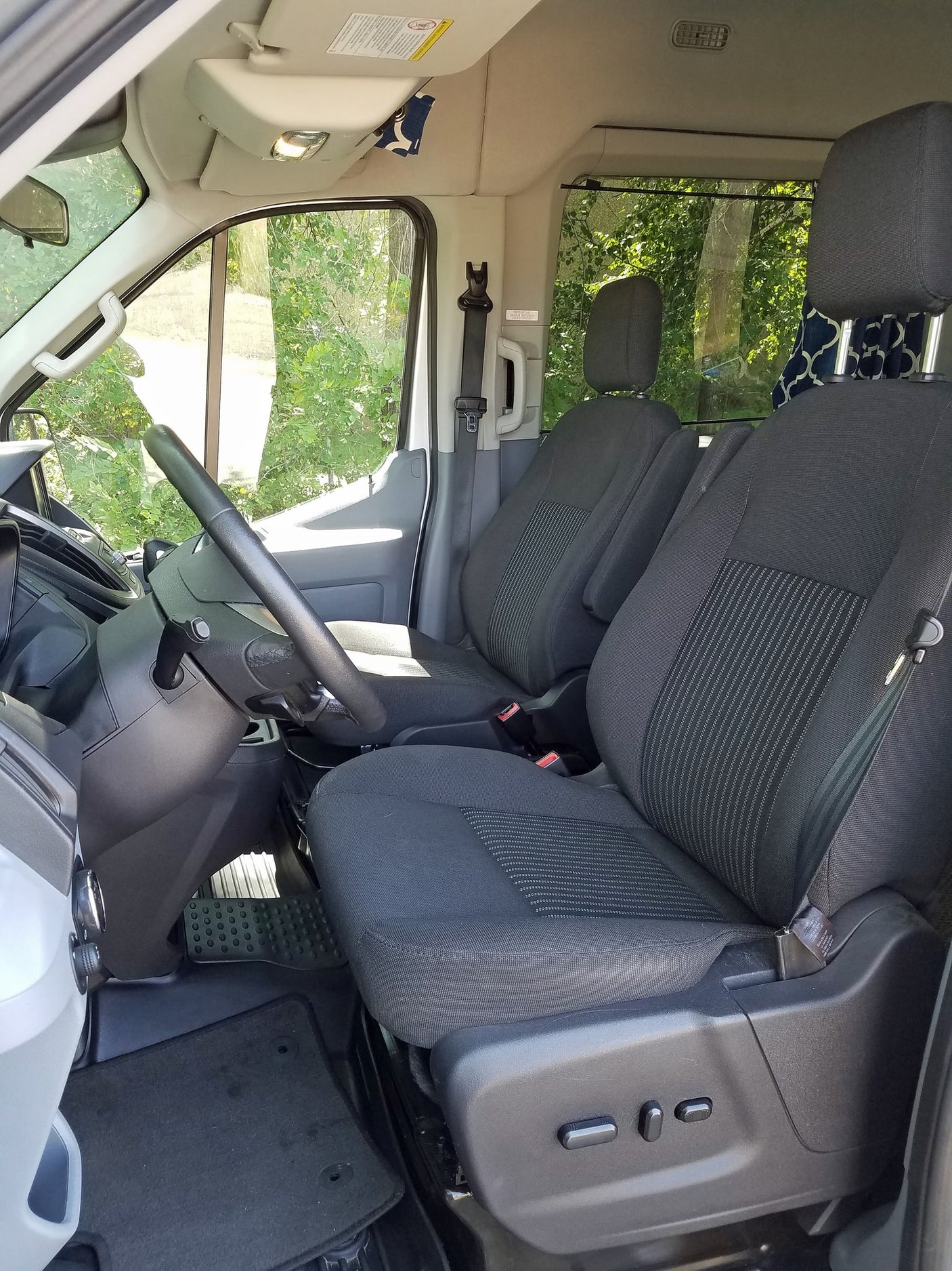 Inside an RV rental: front cabin with dark fabric driver and passenger seats, steering wheel, and power controls. Green trees outside.