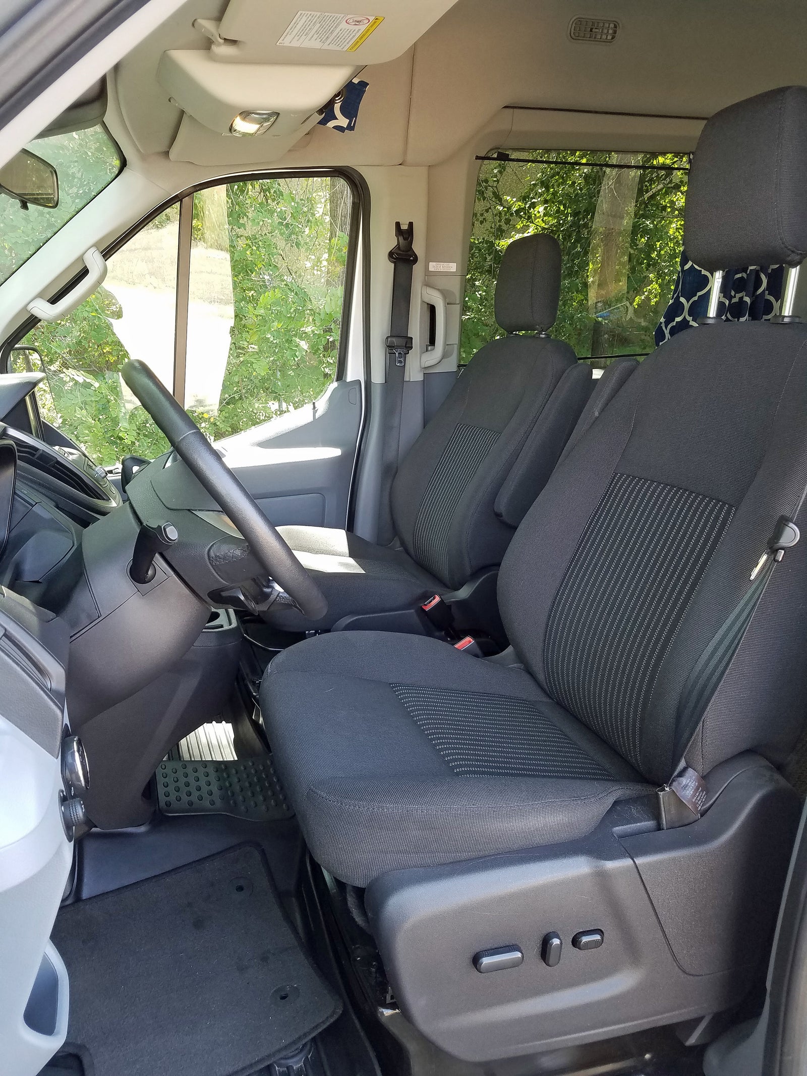 Inside an RV rental: front cabin with dark fabric driver and passenger seats, steering wheel, and power controls. Green trees outside.