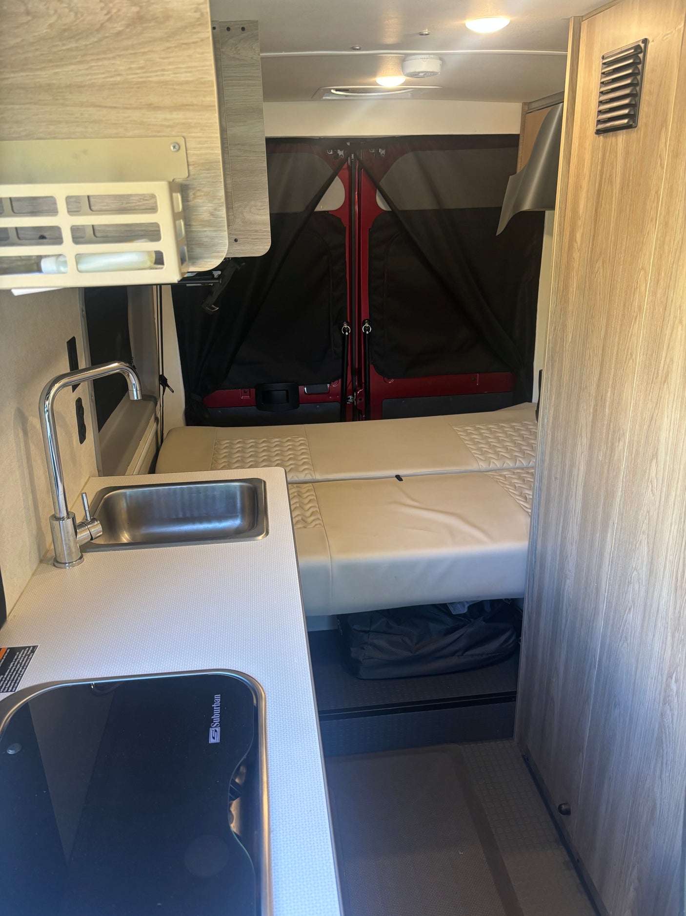 Compact rv rental interior showing kitchenette with sink/stove, bed, overhead cabinets, and rear privacy screens. Ideal for travel.