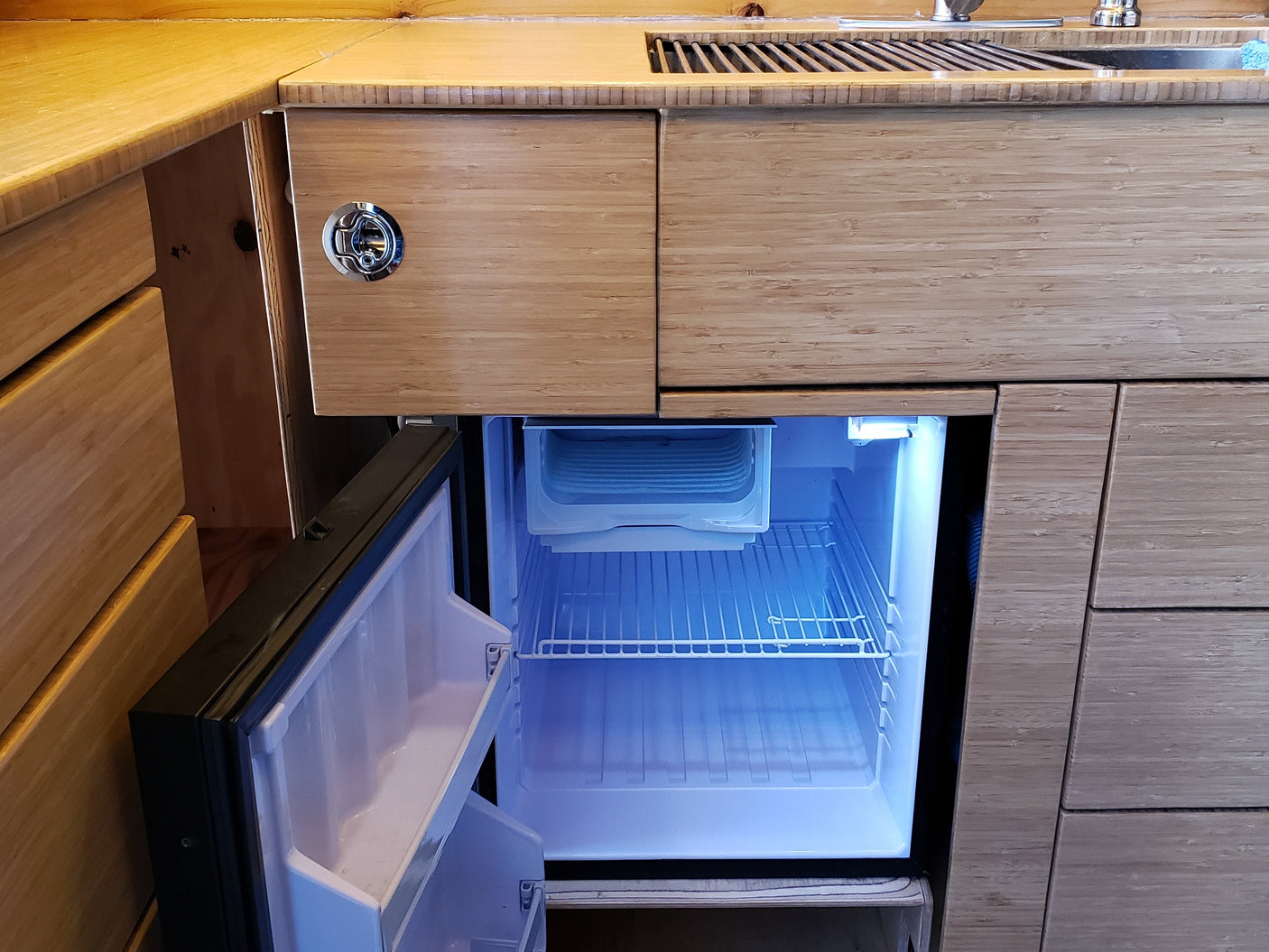 RV rental: Bamboo kitchen counter, sink, open illuminated mini-fridge, and cabinets with a round latch.