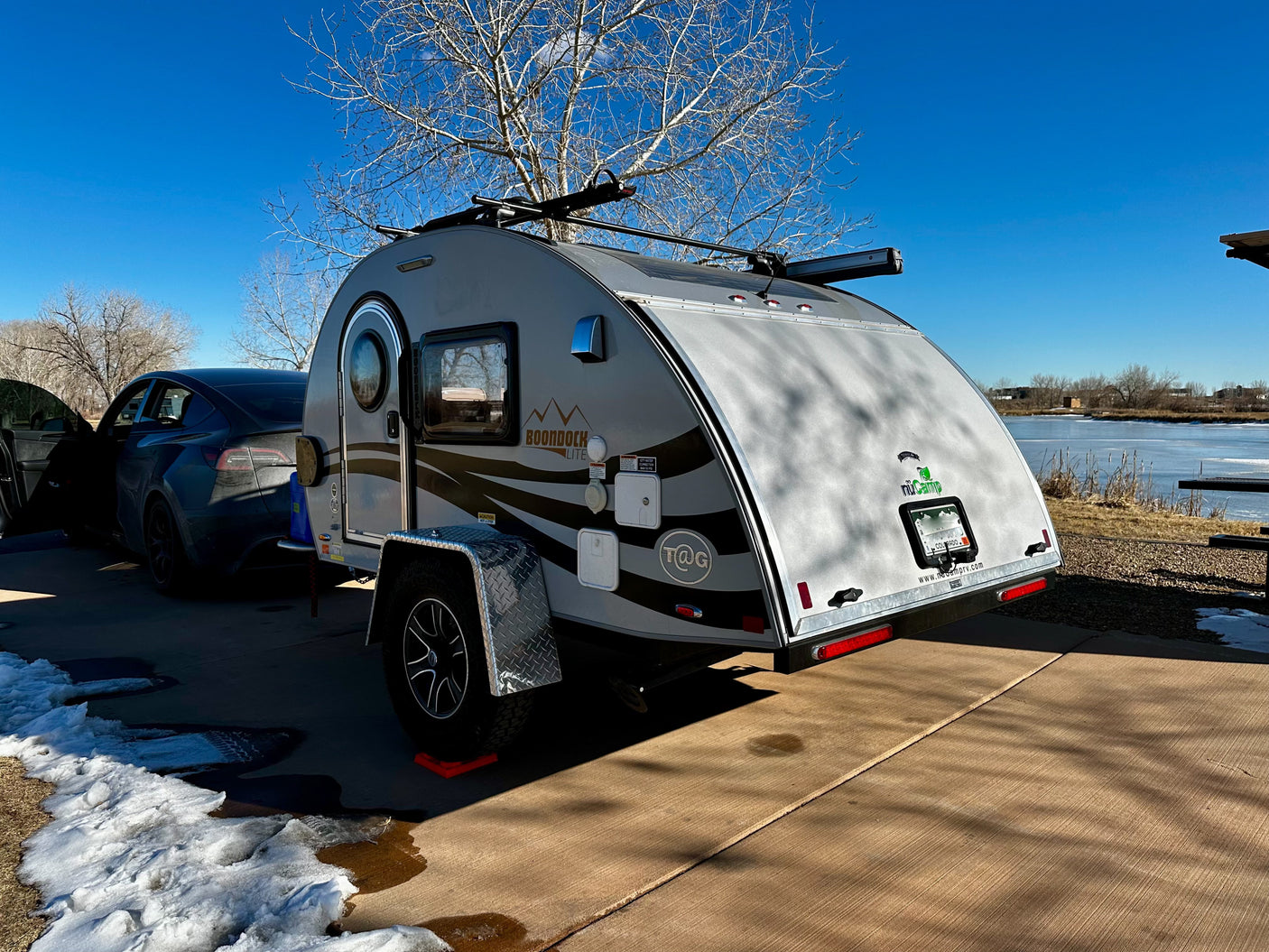 Scenic RV rental spot: nuCamp teardrop camper & Tesla by a frozen lake with patches of snow.