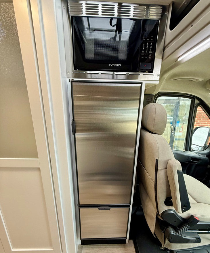 RV rental interior shows a compact stainless steel microwave over a refrigerator, next to a comfy driver's seat.