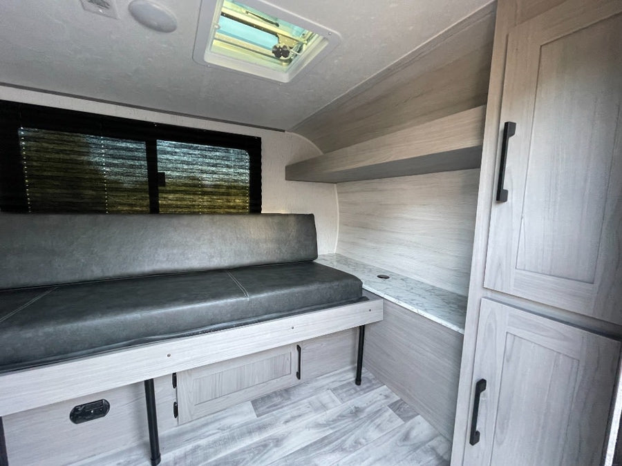 Inside a compact RV rental: gray bench seating, light wood-look interior, storage cabinets, and skylight.