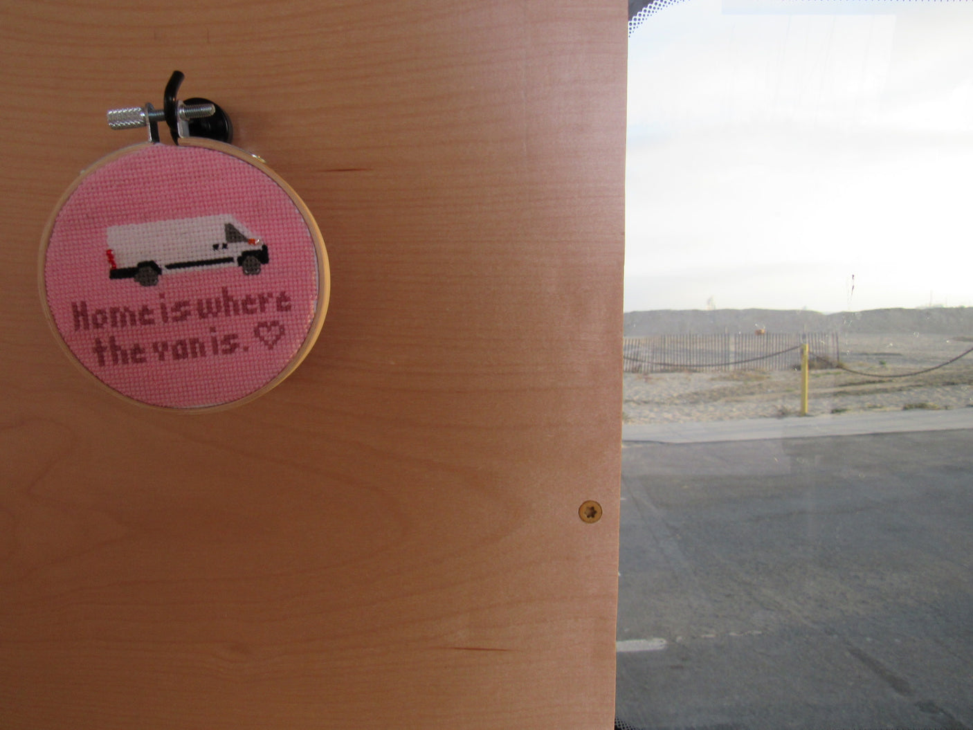 A cross-stitch "Home is where the van is" in an RV rental interior, with a sandy view and road outside. Van life decor.