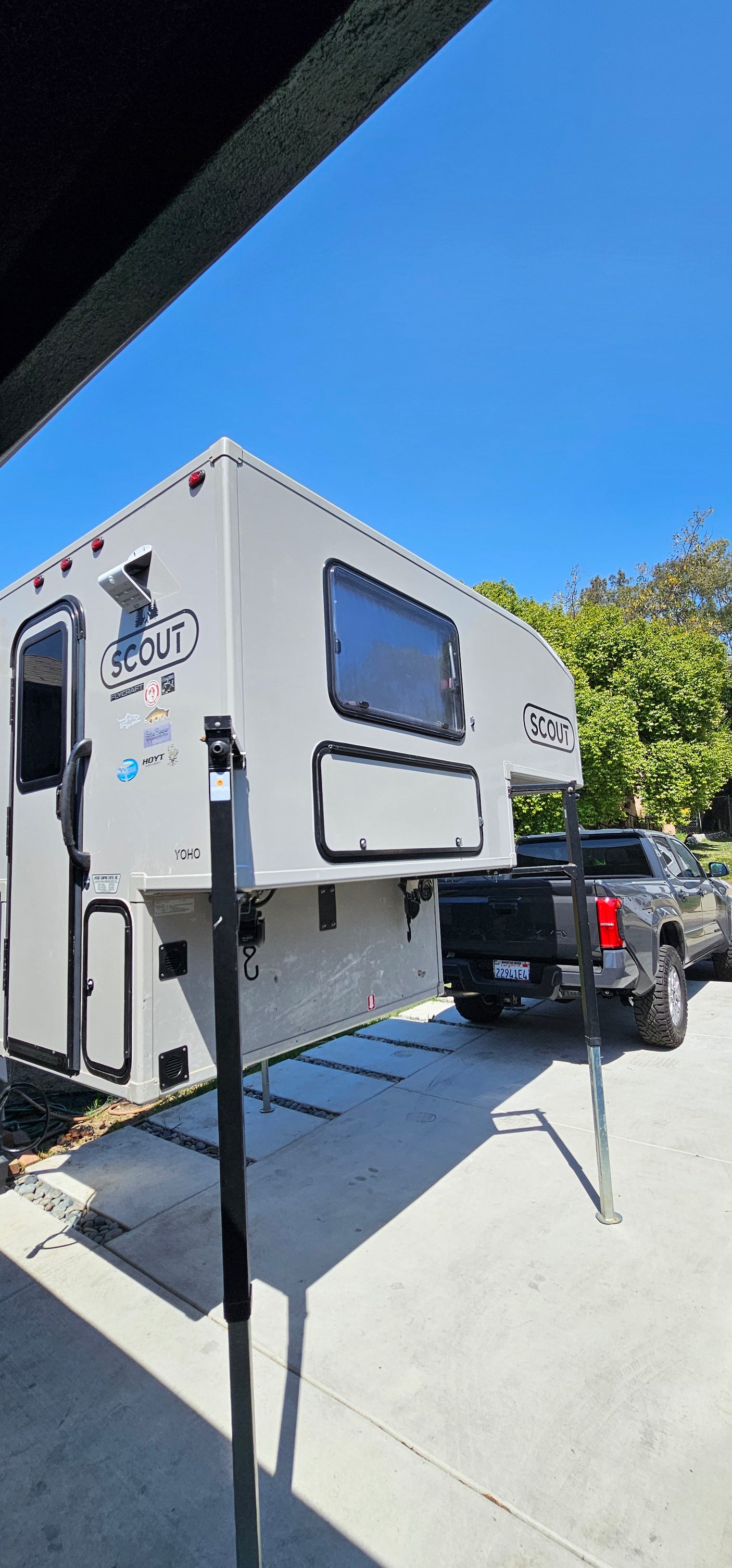 Light gray Scout Yoho truck camper on jacks next to a dark pickup truck on concrete. Perfect for RV rental outdoor adventures.