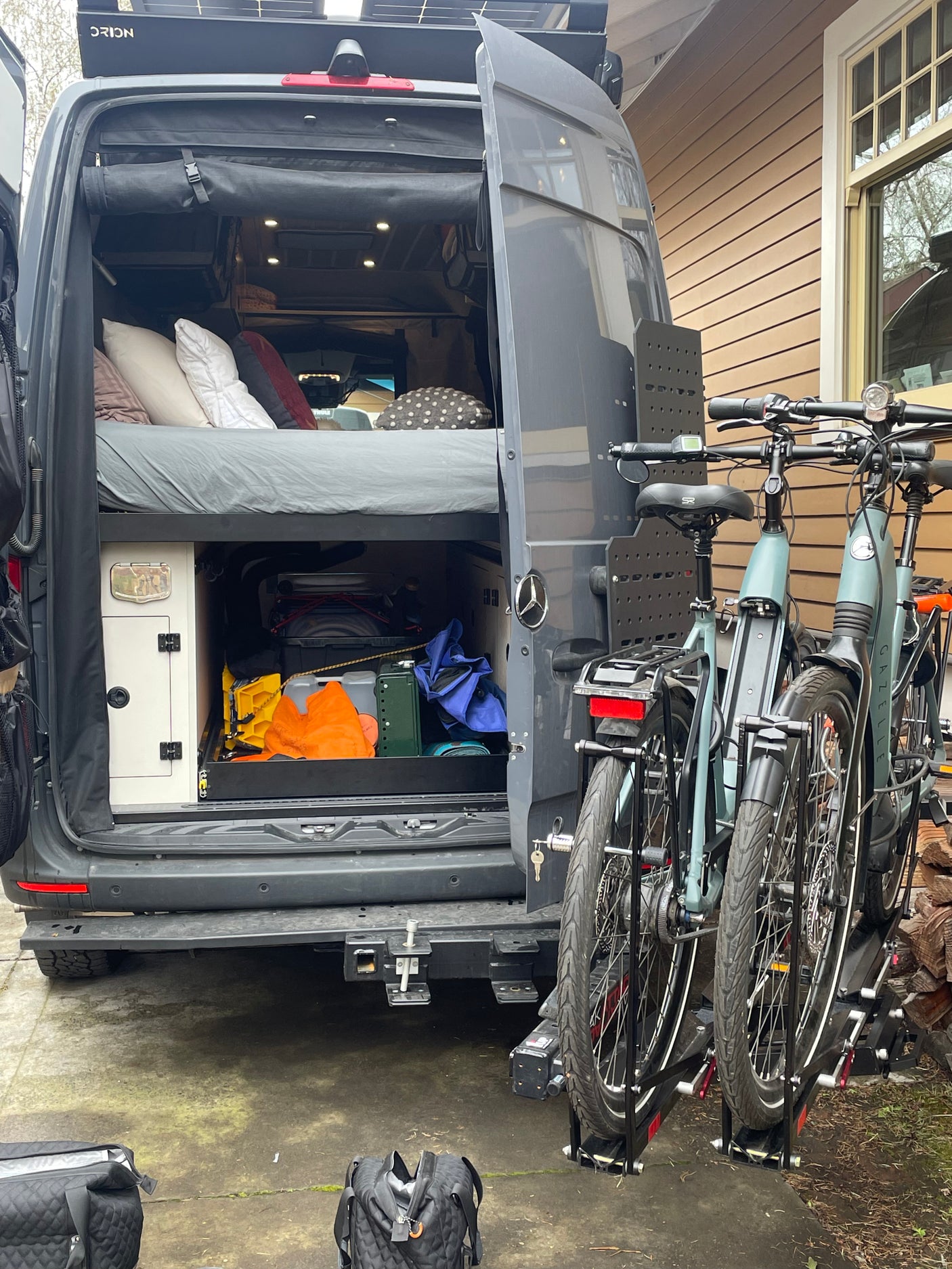 Dark grey camper van with open rear doors showing bed and storage. Roof solar and two e-bikes on rack. Perfect RV rental for adventure!