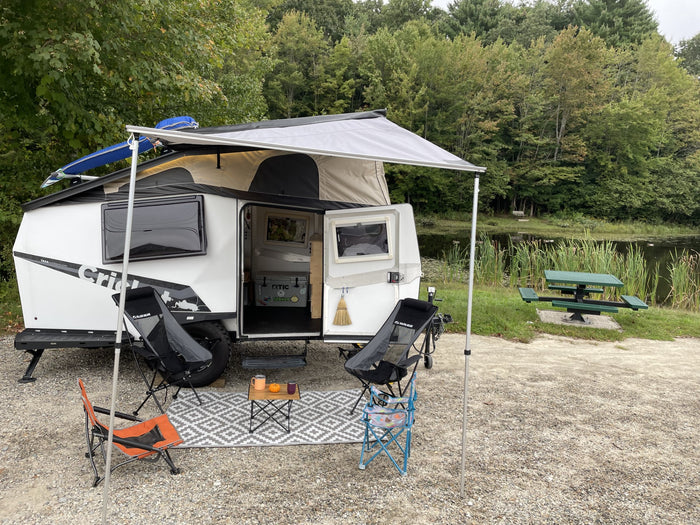 and table are set up by a calm pond.|Lakeside RV rental setup: a white TAXA Cricket camper with a paddleboard on top