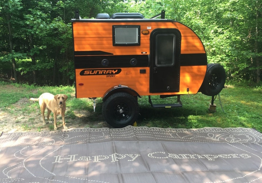 Yellow lab by a vibrant orange Sunray Sport RV rental at a wooded campsite, with a 'Happy Campers' mat. Great for travel!