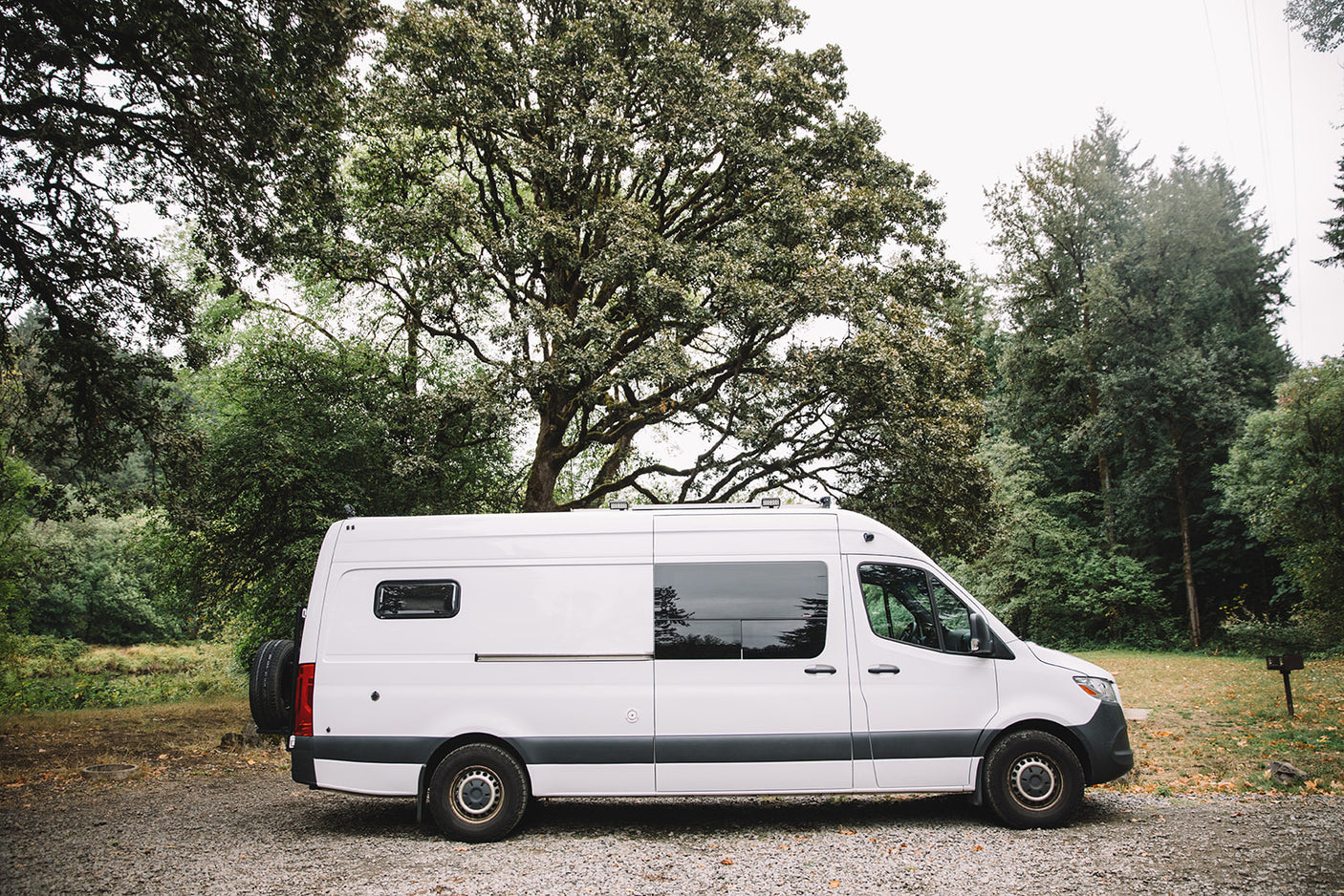 A sleek white Mercedes Sprinter camper van, perfect for your next RV rental adventure, parked on a gravel lot in a wooded area.