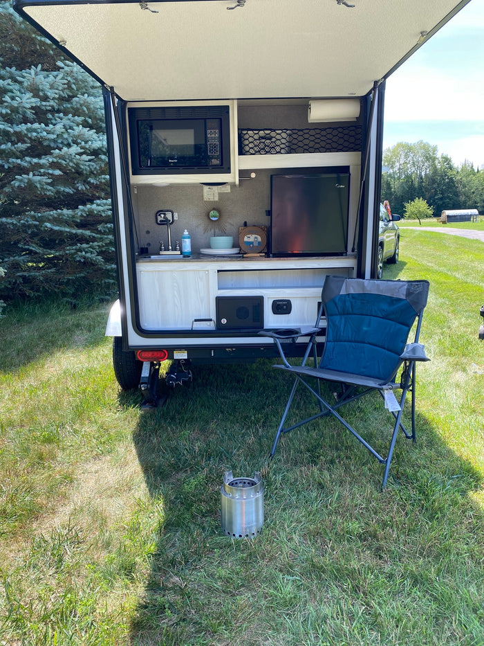 Compact RV rental with rear kitchen (microwave, fridge, sink) open for outdoor use. Camping chair & Solo Stove nearby.