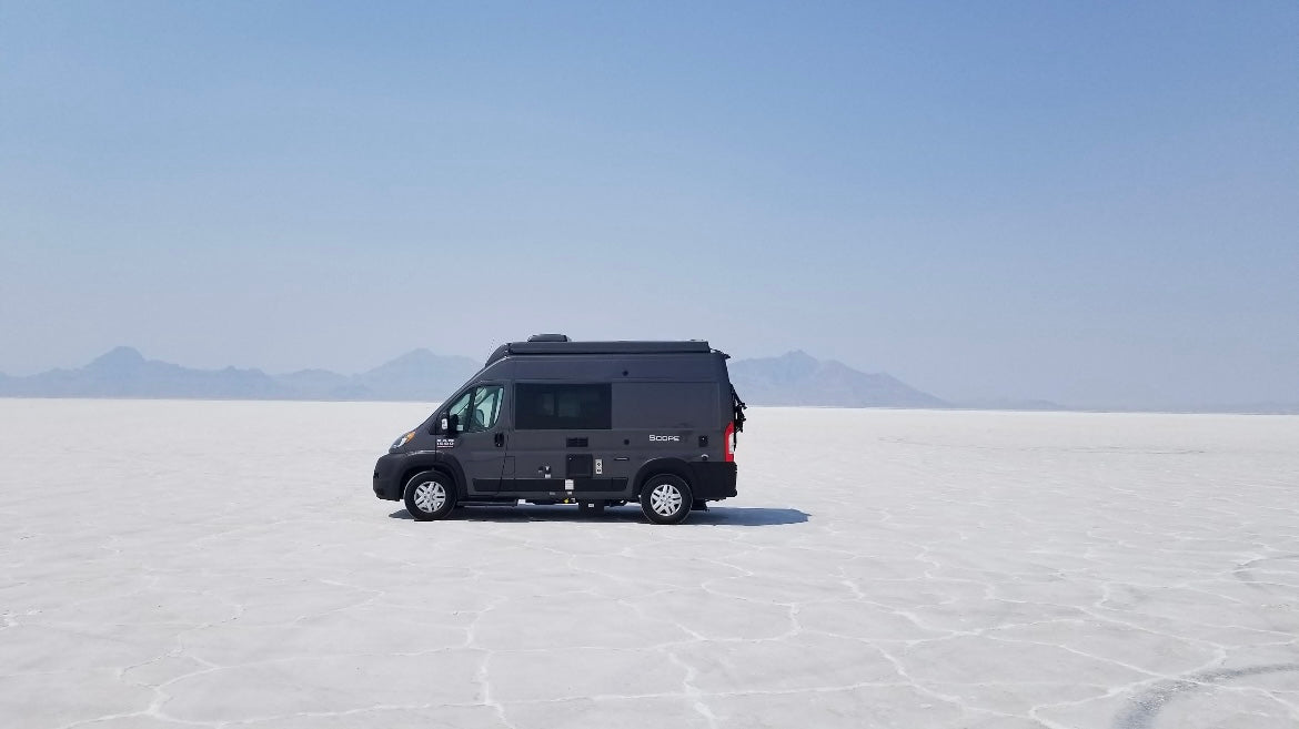 RV rental freedom: Dark grey camper van on vast white Bonneville Salt Flats, mountains under hazy sky.