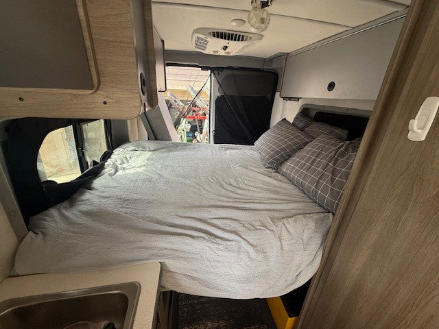 Interior view of an RV rental: a comfortable bed with grey bedding and pillows, and a small sink area.