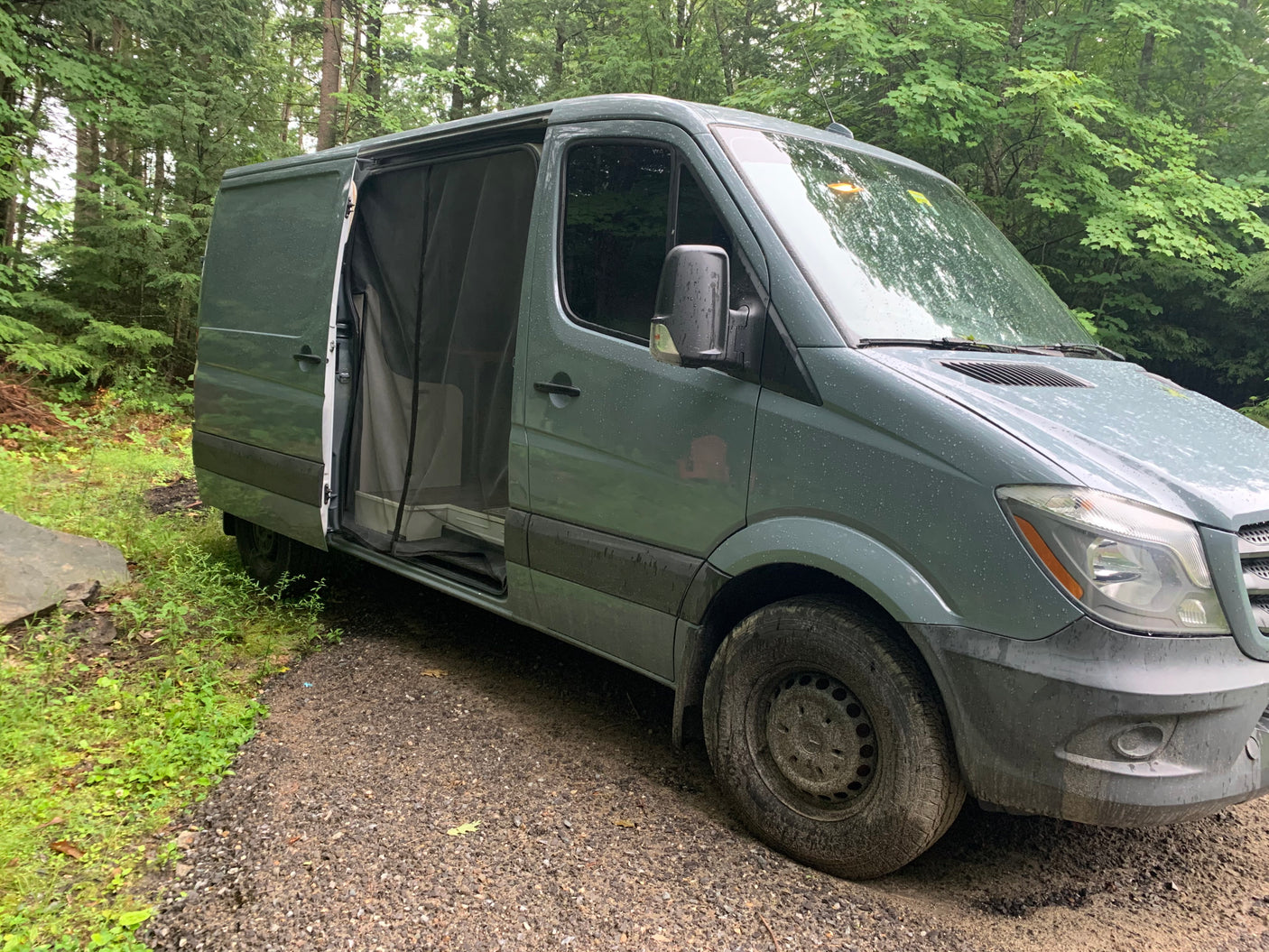 Dark green camper van, likely an rv rental, with its side door open on a gravel path in a lush forest.