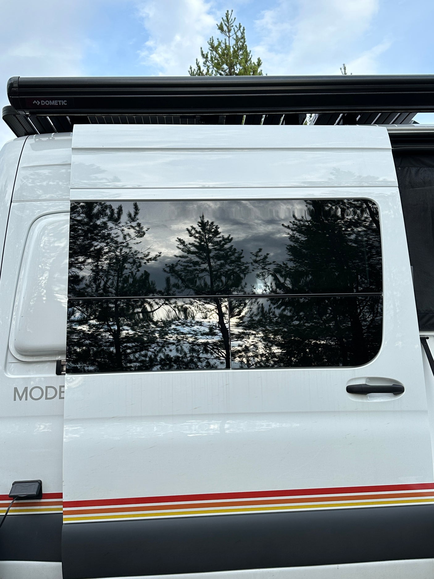 Side exterior of a white rv rental camper van, showing Dometic awning, reflective windows with trees, and colored stripes.