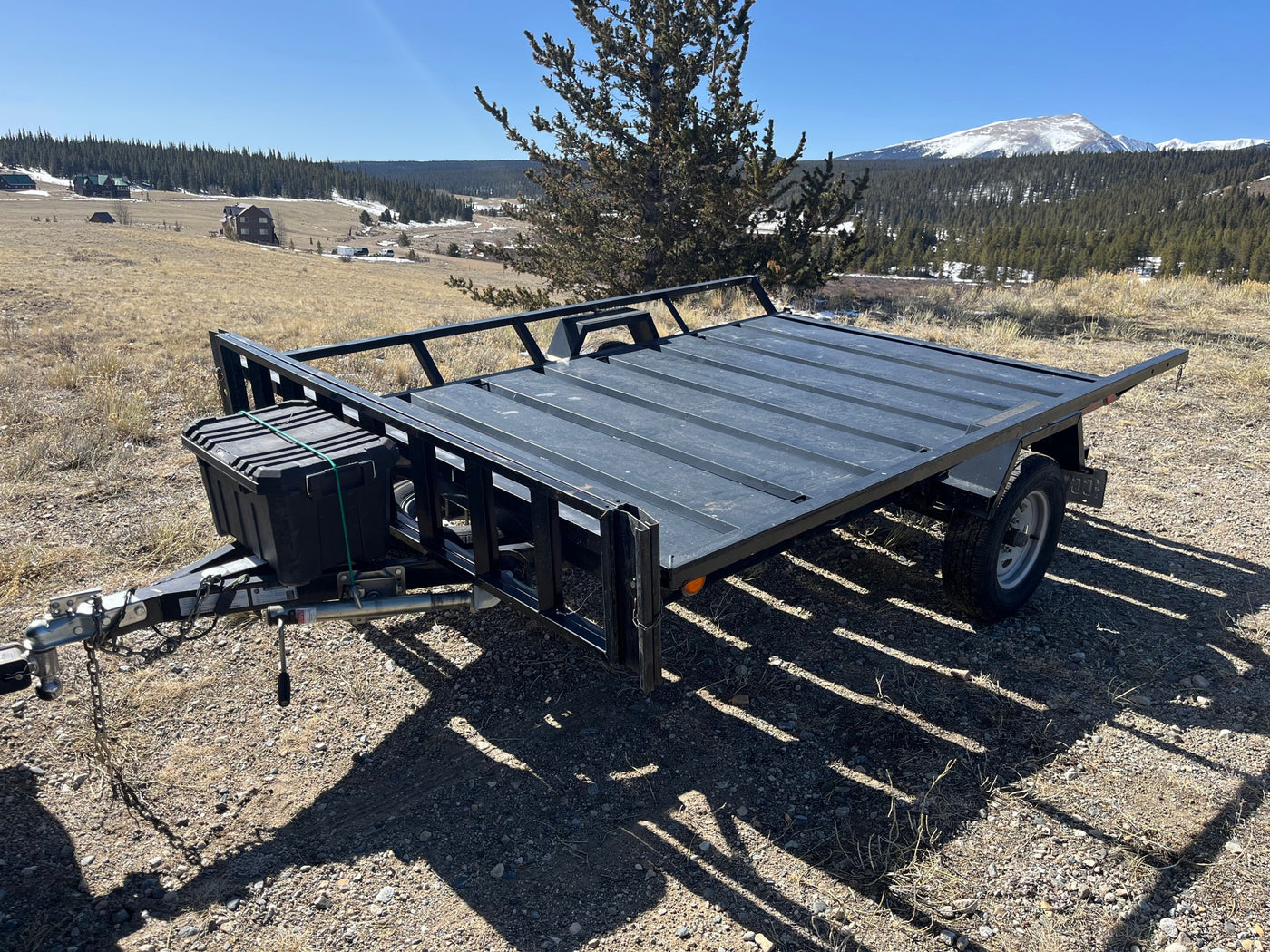 A black utility trailer, ideal for your RV rental, is parked on a dirt hill with scenic mountains and cabins in the distance. — photo 2