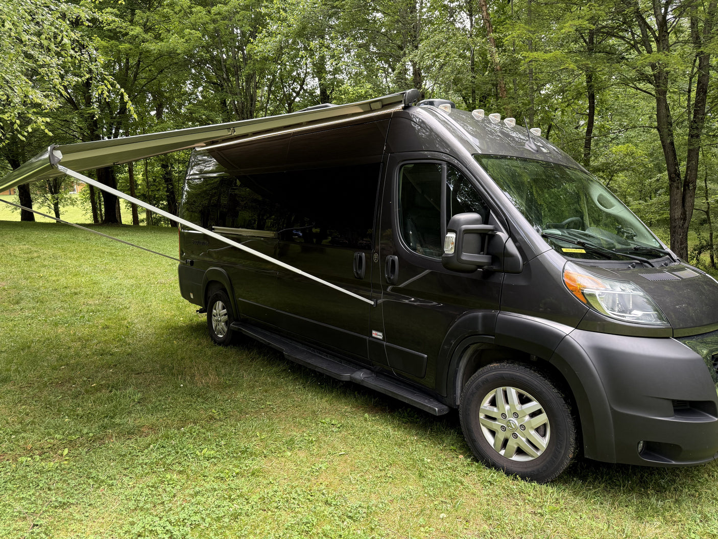 Dark gray campervan with an extended awning parked on green grass amidst trees, perfect for an RV rental getaway.