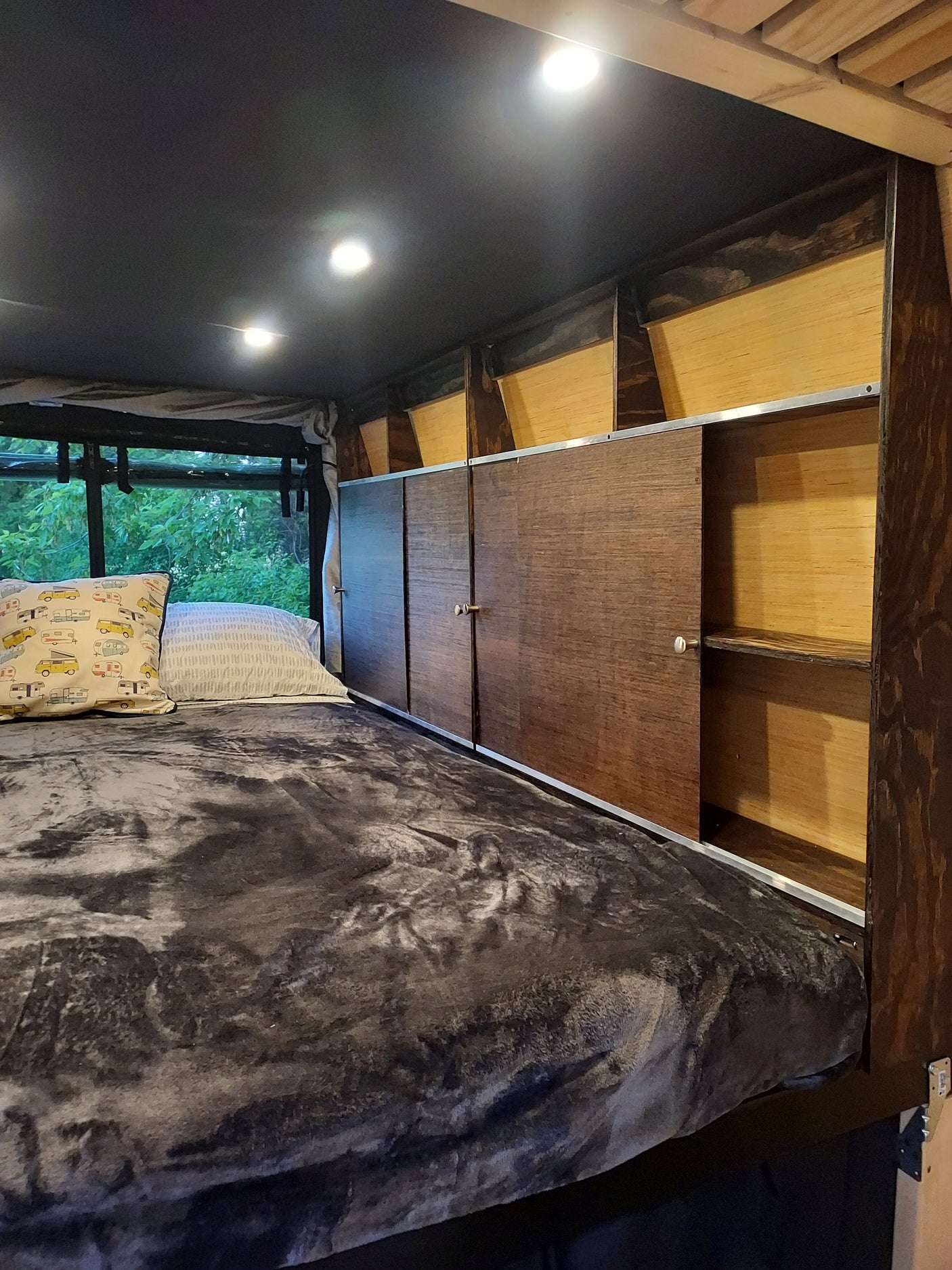 Cozy RV rental bedroom featuring a comfy bed with a plush blanket, bright lights, and overhead paneling with a window view.