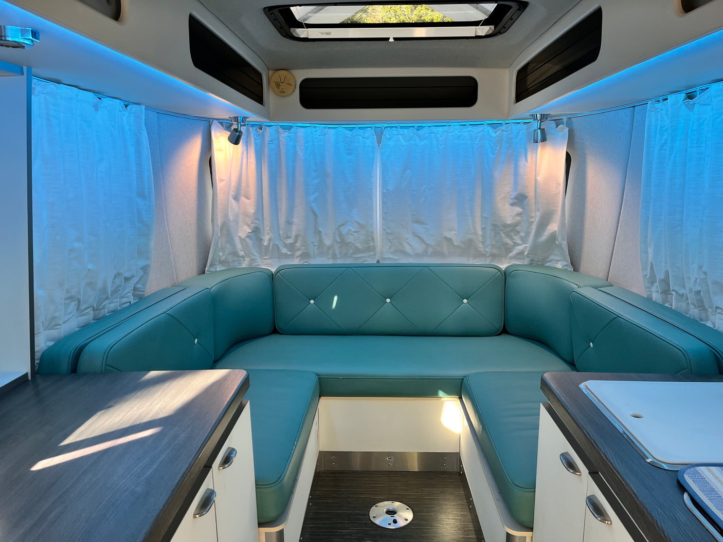 Interior of an rv rental: U-shaped turquoise dinette, ambient blue lighting, overhead storage, and counters. A modern camper layout.