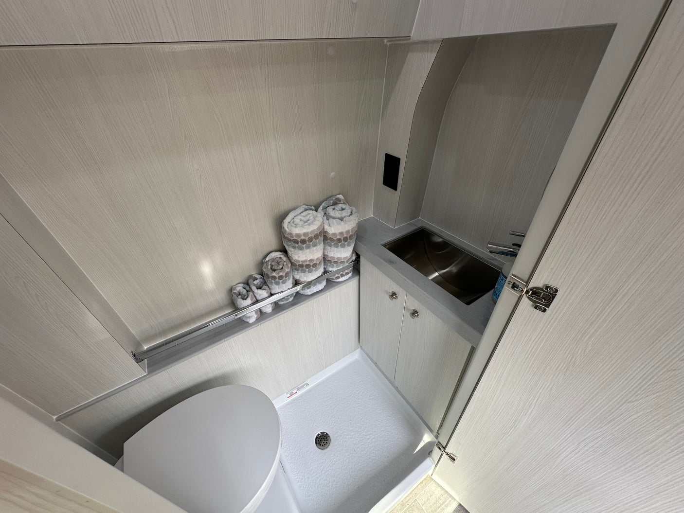 This RV rental features a compact wet bath with a toilet, shower base, stainless steel sink, and light wood-textured interior.